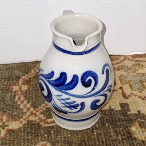 Puede incluir: Una jarra de cerámica blanca con pico y asa, adornada con motivos florales azules. La jarra tiene un cuerpo redondeado y una banda azul alrededor de la parte superior. Esta jarra decorativa es adecuada para servir bebidas.