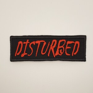 May include: Black and red embroidered patch with the word "DISTURBED" in red lettering.