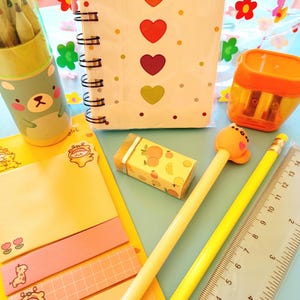 May include: A collection of stationery items, including a spiral-bound notebook with heart designs, pencils, a pencil sharpener, sticky notes, a ruler, and an eraser. The items are in various colors, including yellow, orange, and green, and are arranged on a light blue surface.