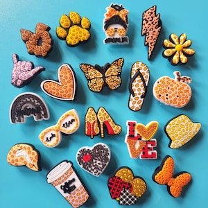 May include: A collection of decorative shoe charms in various shapes and designs. Includes a paw print, butterfly, high heels, coffee cup, and a lightning bolt. The charms feature patterns like leopard print, plaid, and floral designs. The background is a solid light blue.