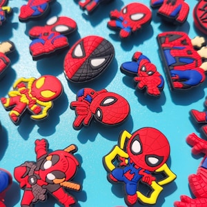 Puede incluir: Una colecci&oacute;n de dijes para zapatos del personaje de Spider-Man en rojo, azul y negro. Los dijes presentan varias poses y dise&ntilde;os del superh&eacute;roe, algunos con detalles amarillos. El fondo es azul claro.