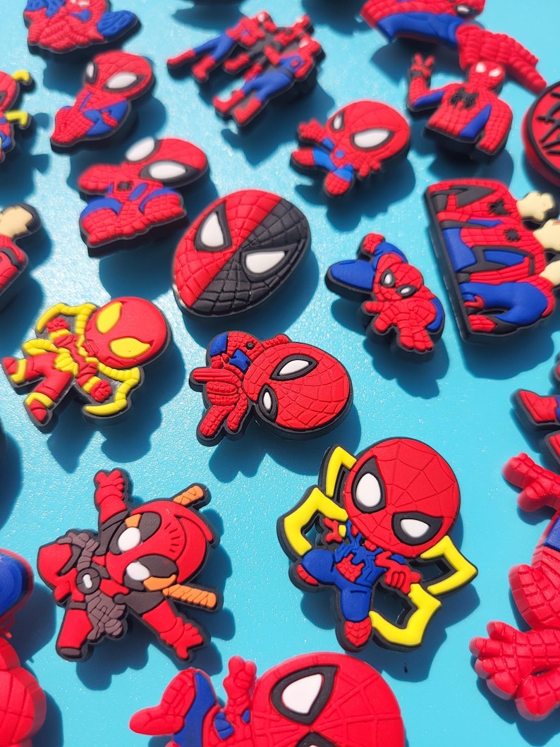 Puede incluir: Una colecci&oacute;n de dijes para zapatos del personaje de Spider-Man en rojo, azul y negro. Los dijes presentan varias poses y dise&ntilde;os del superh&eacute;roe, algunos con detalles amarillos. El fondo es azul claro.