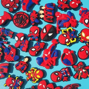Puede incluir: Una colecci&oacute;n de dijes y adornos con tem&aacute;tica de Spider-Man en rojo, azul y negro. Los dijes presentan varias poses y dise&ntilde;os del superh&eacute;roe, incluyendo su m&aacute;scara, logotipo y figuras de cuerpo completo. El fondo es de un azul claro s&oacute;lido.