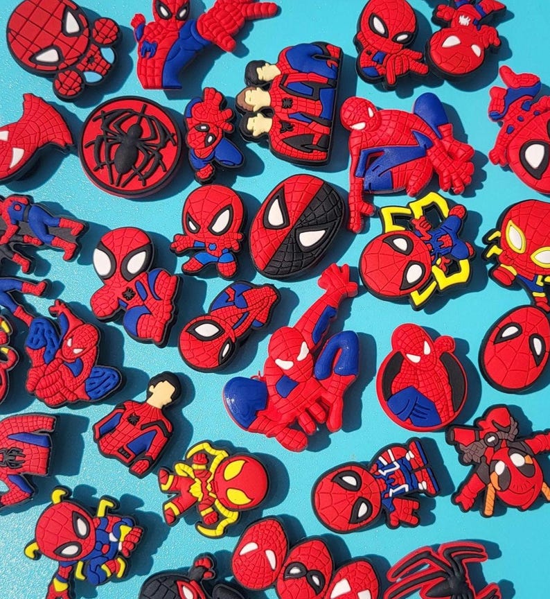 Puede incluir: Una colecci&oacute;n de dijes y adornos con tem&aacute;tica de Spider-Man en rojo, azul y negro. Los dijes presentan varias poses y dise&ntilde;os del superh&eacute;roe, incluyendo su m&aacute;scara, logotipo y figuras de cuerpo completo. El fondo es de un azul claro s&oacute;lido.