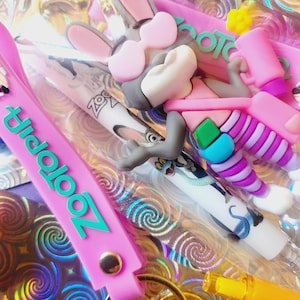 Puede incluir: Una colecci&oacute;n de accesorios con tem&aacute;tica de Zootopia, que incluye un llavero rosa con la palabra "Zootopia" en turquesa, un bol&iacute;grafo con Judy Hopps y una figura 3D de Nick Wilde con gafas de sol. Los art&iacute;culos est&aacute;n sobre un fondo hologr&aacute;fico.