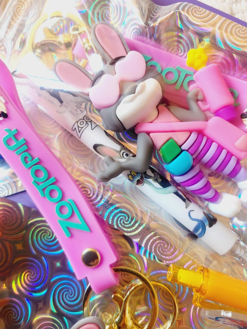 Puede incluir: Una colecci&oacute;n de accesorios con tem&aacute;tica de Zootopia, que incluye un llavero rosa con la palabra "Zootopia" en turquesa, un bol&iacute;grafo con Judy Hopps y una figura 3D de Nick Wilde con gafas de sol. Los art&iacute;culos est&aacute;n sobre un fondo hologr&aacute;fico.