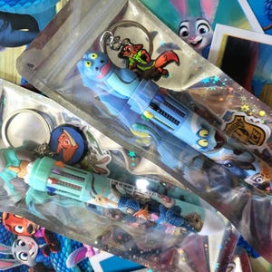 Pu&ograve; includere: Diverse penne e portachiavi a tema Zootropolis in confezioni di plastica trasparente. Le penne sono blu e verde acqua con disegni di personaggi. I portachiavi presentano personaggi dei cartoni animati.