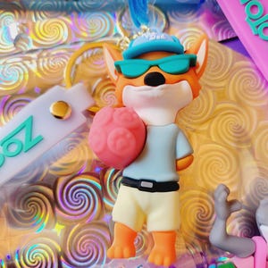 Puede incluir: Un llavero tem&aacute;tico de Zootopia con un personaje de zorro de dibujos animados que lleva gafas de sol y un sombrero, sosteniendo un objeto rosa con forma de cerebro. El zorro viste una camisa azul claro y pantalones cortos amarillos. El fondo tiene un patr&oacute;n de remolino.