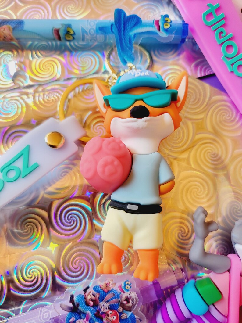 Puede incluir: Un llavero tem&aacute;tico de Zootopia con un personaje de zorro de dibujos animados que lleva gafas de sol y un sombrero, sosteniendo un objeto rosa con forma de cerebro. El zorro viste una camisa azul claro y pantalones cortos amarillos. El fondo tiene un patr&oacute;n de remolino.