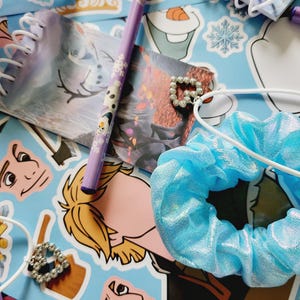 Peut inclure: Une collection de papeterie et d'accessoires comprenant un chouchou bleu clair m&eacute;tallis&eacute;, un crayon violet avec des graphismes de personnages, deux petits carnets &agrave; spirales, deux breloques en forme de c&oelig;ur avec des perles et divers autocollants.