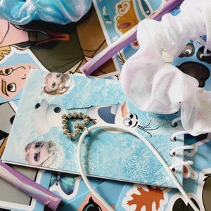 Peut inclure: Un chouchou bleu clair m&eacute;tallis&eacute;, un chouchou blanc transparent et un collier avec un pendentif c&oelig;ur dor&eacute; pos&eacute;s sur un carnet &agrave; spirales illustr&eacute; de personnages de dessins anim&eacute;s et un stylo violet clair sur des autocollants.