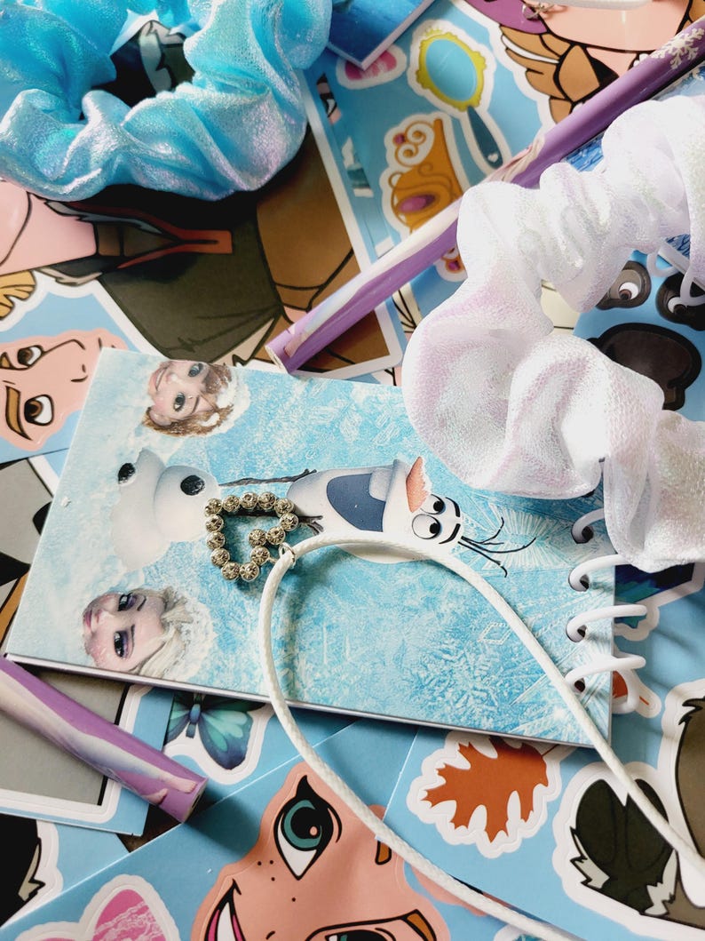 Peut inclure: Un chouchou bleu clair m&eacute;tallis&eacute;, un chouchou blanc transparent et un collier avec un pendentif c&oelig;ur dor&eacute; pos&eacute;s sur un carnet &agrave; spirales illustr&eacute; de personnages de dessins anim&eacute;s et un stylo violet clair sur des autocollants.