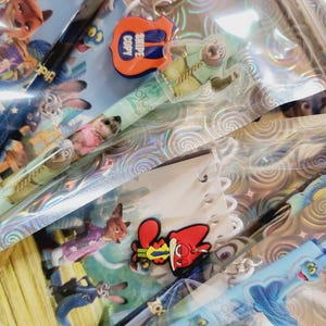 May include: A selection of Zootropolis-themed pens and charms. The pens feature character designs and are in clear plastic packaging. Charms include a guitar-shaped charm with the text "SNIPE COPY". The background has a holographic pattern.