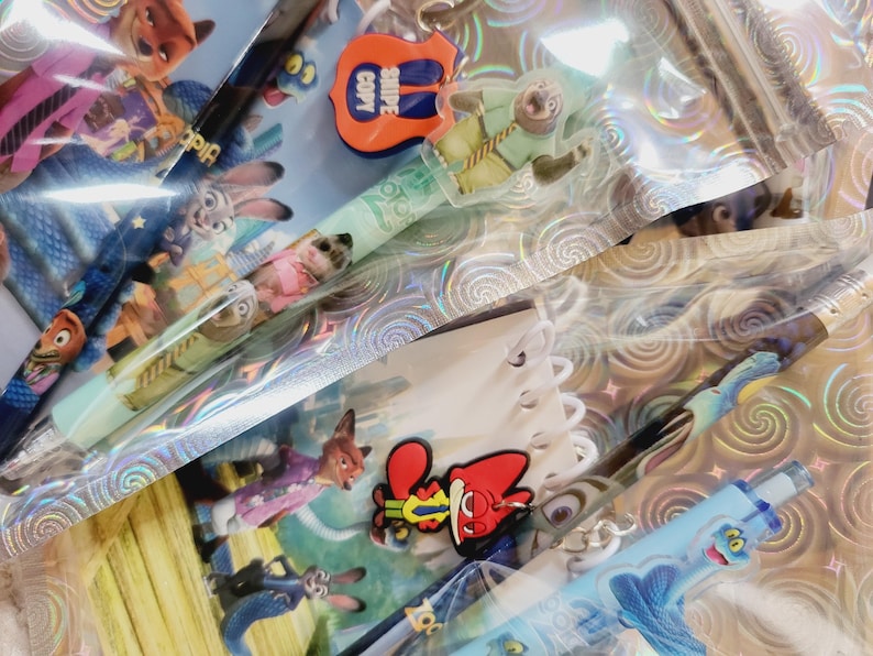 May include: A selection of Zootropolis-themed pens and charms. The pens feature character designs and are in clear plastic packaging. Charms include a guitar-shaped charm with the text "SNIPE COPY". The background has a holographic pattern.