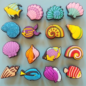 Könnte beinhalten: Eine Sammlung farbenfroher, cartoonartiger Meereslebewesen-Anhänger, darunter Fische und Muscheln. Die Anhänger weisen leuchtende Farben wie Gelb, Blau, Rosa und Grün mit schwarzen Umrissen auf. Die Designs sind detailliert und skurril.