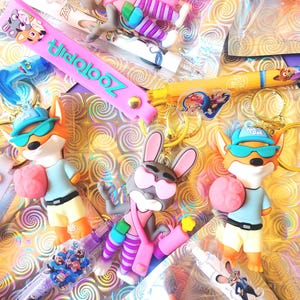 Puede incluir: Una colecci&oacute;n de productos con tem&aacute;tica de Zootopia, que incluye llaveros, bol&iacute;grafos y pulseras. Los llaveros presentan personajes de dibujos animados como un zorro y un conejo. La pulsera es rosa con la palabra "Zootopia" en turquesa. Los bol&iacute;grafos tienen dise&ntilde;os de personajes.