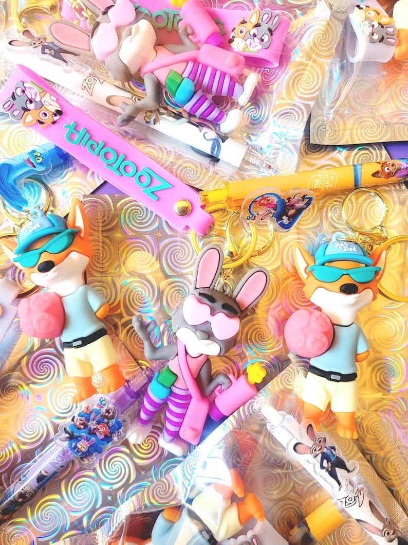 Puede incluir: Una colecci&oacute;n de productos con tem&aacute;tica de Zootopia, que incluye llaveros, bol&iacute;grafos y pulseras. Los llaveros presentan personajes de dibujos animados como un zorro y un conejo. La pulsera es rosa con la palabra "Zootopia" en turquesa. Los bol&iacute;grafos tienen dise&ntilde;os de personajes.