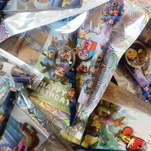 May include: A collection of Zootropolis-themed stationery and accessories, including pens, keyrings, and stickers. The items are packaged in clear, iridescent cellophane. A keyring features Judy Hopps and Nick Wilde. Some items have "Happy Birthday" text.