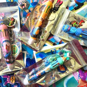 Pu&ograve; includere: Diverse penne multicolore con personaggi del film d'animazione "Zootropolis" sono esposte in confezioni di plastica trasparente. Ogni penna ha un personaggio diverso in cima e un portachiavi abbinato. Le penne sono in vari colori, tra cui rosa, blu e giallo.