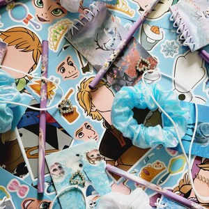 Peut inclure: Une collection de papeterie et d'accessoires sur le th&egrave;me de La Reine des Neiges comprenant des crayons violets &agrave; motifs de flocons, des petits carnets &agrave; spirales, des chouchous en tissu bleu et blanc, et des breloques en forme de c&oelig;ur.