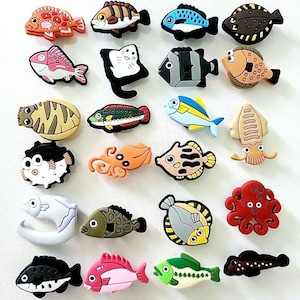 Könnte beinhalten: Eine Sammlung farbenfroher, cartoonartiger Fisch- und Meereskreaturen-Anhänger. Die Anhänger weisen verschiedene Designs auf, darunter ein roter Oktopus, ein blau-gelber Fisch und ein schwarz-weißer Fisch. Diese Anhänger dienen wahrscheinlich dekorativen Zwecken.