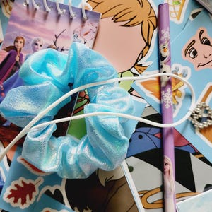 Peut inclure: Un chouchou en tissu bleu clair m&eacute;tallis&eacute; avec un cordon &eacute;lastique blanc, pos&eacute; sur une collection d'autocollants et de papeterie repr&eacute;sentant des personnages et motifs du film La Reine des Neiges, incluant un carnet &agrave; spirales et un crayon violet.