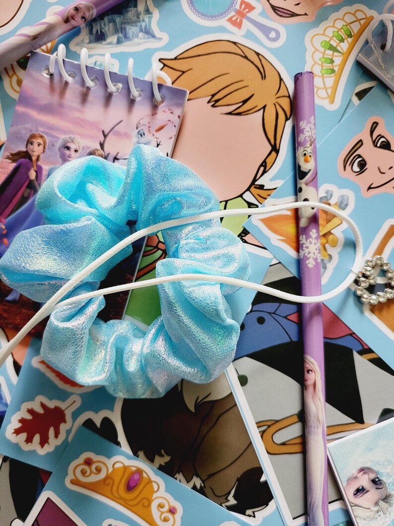 Peut inclure: Un chouchou en tissu bleu clair m&eacute;tallis&eacute; avec un cordon &eacute;lastique blanc, pos&eacute; sur une collection d'autocollants et de papeterie repr&eacute;sentant des personnages et motifs du film La Reine des Neiges, incluant un carnet &agrave; spirales et un crayon violet.