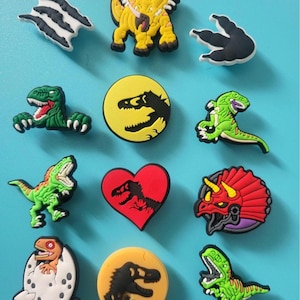 Könnte beinhalten: Eine Sammlung bunter Schuhanhänger zum Thema Dinosaurier. Die Anhänger zeigen verschiedene Dinosaurier-Designs, darunter einen T-Rex, Triceratops und Dinosaurier-Fußabdrücke. Die Anhänger sind in den Farbtönen Grün, Gelb, Rot und Schwarz gehalten, einige in Herz- und Eiform.