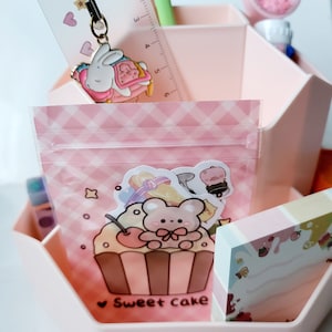 Puede incluir: Un organizador de escritorio hexagonal rosa con una bolsa de plástico transparente que contiene una pegatina de un cupcake con un oso en la parte superior. La pegatina dice "Sweet Cake". Hay otras pegatinas en la bolsa, incluyendo un cerdo y un gato. El organizador tiene una regla, un bolígrafo y un pequeño recipiente rosa con purpurina en su interior.