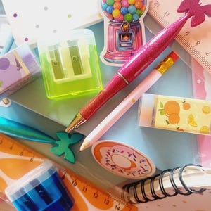 May include: A collection of stationery items including a pink pen, a pencil, a blue and green pencil sharpener, a blue pencil sharpener, a ruler with a citrus design, a donut sticker, a gumball machine sticker, a butterfly sticker, and a small notebook with a white and pink polka dot design.