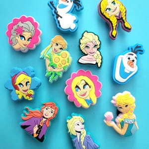 Può includere: Una collezione di ciondoli colorati per scarpe con personaggi dei cartoni animati. I ciondoli includono immagini di un pupazzo di neve, principesse e altri personaggi con accenti rosa, blu, gialli e viola. I ciondoli sono progettati per essere attaccati alle scarpe.