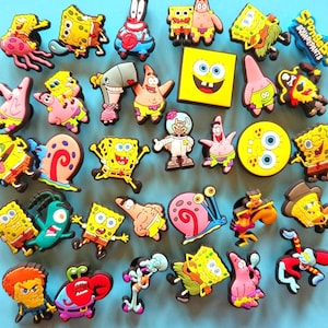 Op de afbeelding: Een verzameling kleurrijke bedels met SpongeBob SquarePants-personages. De bedels tonen verschillende personages zoals SpongeBob, Patrick Star en Octo Tentakel, in heldere gele, roze en blauwe tinten. De bedels zijn gerangschikt op een lichtblauwe achtergrond.
