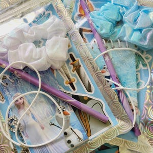 Peut inclure: Deux pochettes en plastique transparent contenant des kits de papeterie avec Elsa et Olaf de La Reine des Neiges. Chaque kit comprend un chouchou iris&eacute; blanc ou bleu clair, un crayon violet &agrave; motifs flocons, un collier cordon blanc et des autocollants.