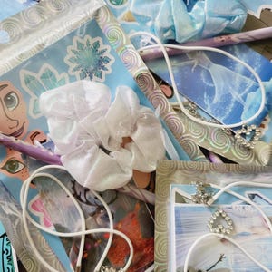 Peut inclure: Un ensemble d'accessoires comprenant deux chouchous en tissu blanc et bleu clair, deux colliers &agrave; cordon blanc avec des pendentifs en forme de c&oelig;ur orn&eacute;s de strass, ainsi que divers autocollants et cartes illustrant des personnages et flocons.