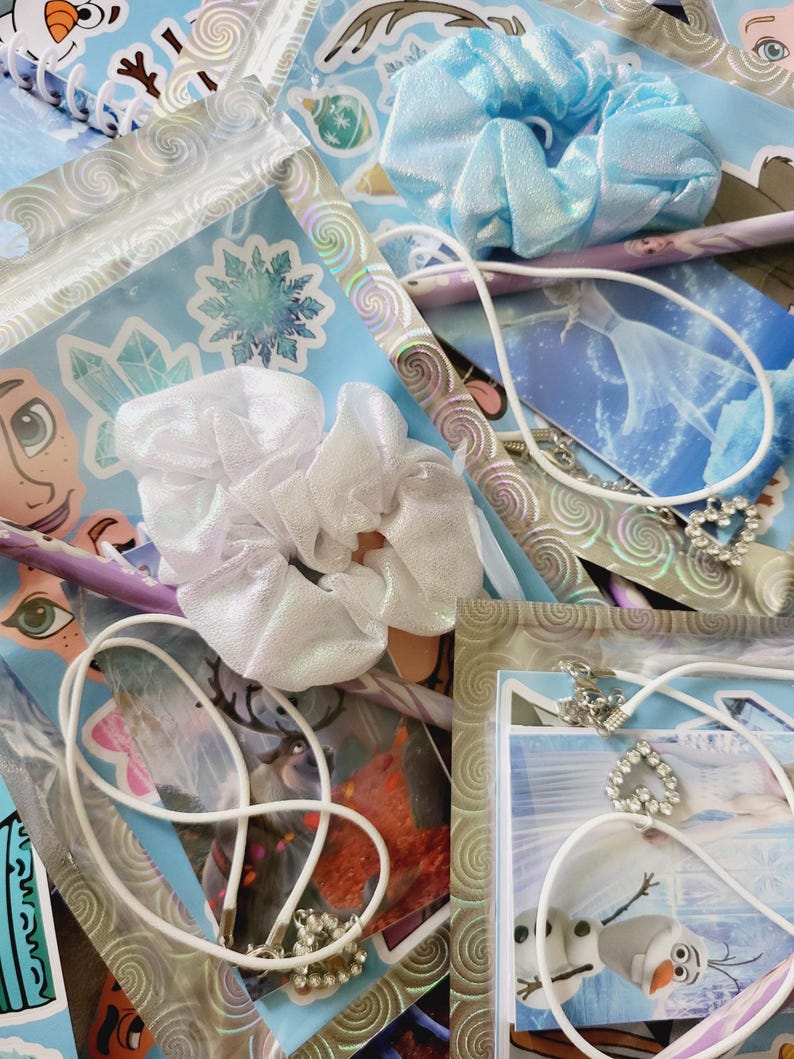 Peut inclure: Un ensemble d'accessoires comprenant deux chouchous en tissu blanc et bleu clair, deux colliers &agrave; cordon blanc avec des pendentifs en forme de c&oelig;ur orn&eacute;s de strass, ainsi que divers autocollants et cartes illustrant des personnages et flocons.