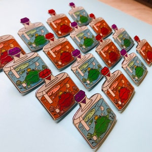 Puede incluir: Una colecci&oacute;n de 12 pines de esmalte que representan una rana y un sapo en una botella de vidrio con un tap&oacute;n de joya. Las botellas est&aacute;n llenas de agua y burbujas, y las ranas llevan coronas. Los pines son de varios colores, incluyendo azul, verde, rojo, naranja y morado.
