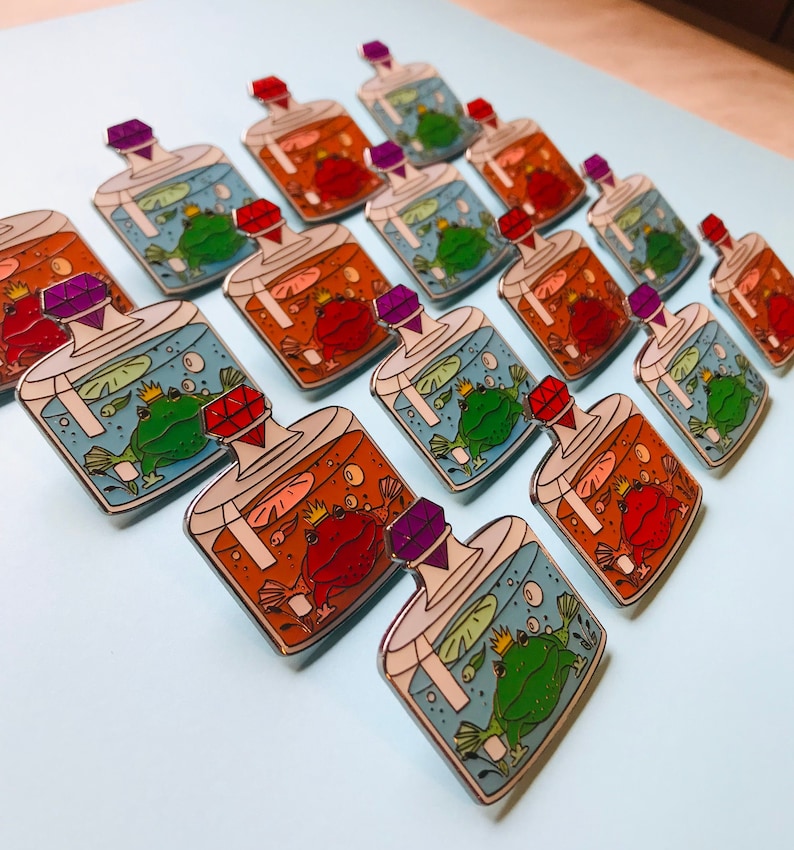 Puede incluir: Una colecci&oacute;n de 12 pines de esmalte que representan una rana y un sapo en una botella de vidrio con un tap&oacute;n de joya. Las botellas est&aacute;n llenas de agua y burbujas, y las ranas llevan coronas. Los pines son de varios colores, incluyendo azul, verde, rojo, naranja y morado.