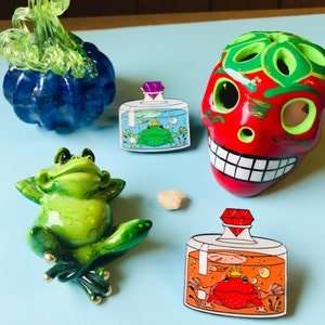 Puede incluir: Dos pines de esmalte que representan frascos de vidrio con ranas en su interior. El primer frasco tiene una rana verde con una corona y una gema p&uacute;rpura en la tapa. El segundo frasco tiene una rana roja con una corona y una gema roja en la tapa.