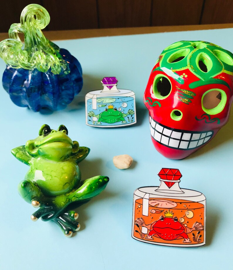 Puede incluir: Dos pines de esmalte que representan frascos de vidrio con ranas en su interior. El primer frasco tiene una rana verde con una corona y una gema p&uacute;rpura en la tapa. El segundo frasco tiene una rana roja con una corona y una gema roja en la tapa.