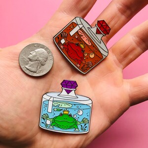 Puede incluir: Dos pines de esmalte que representan ranas en frascos de vidrio con tapas de joyas. El primer pin presenta una rana roja en un frasco naranja con una joya roja. El segundo pin presenta una rana verde en un frasco azul con una joya morada.