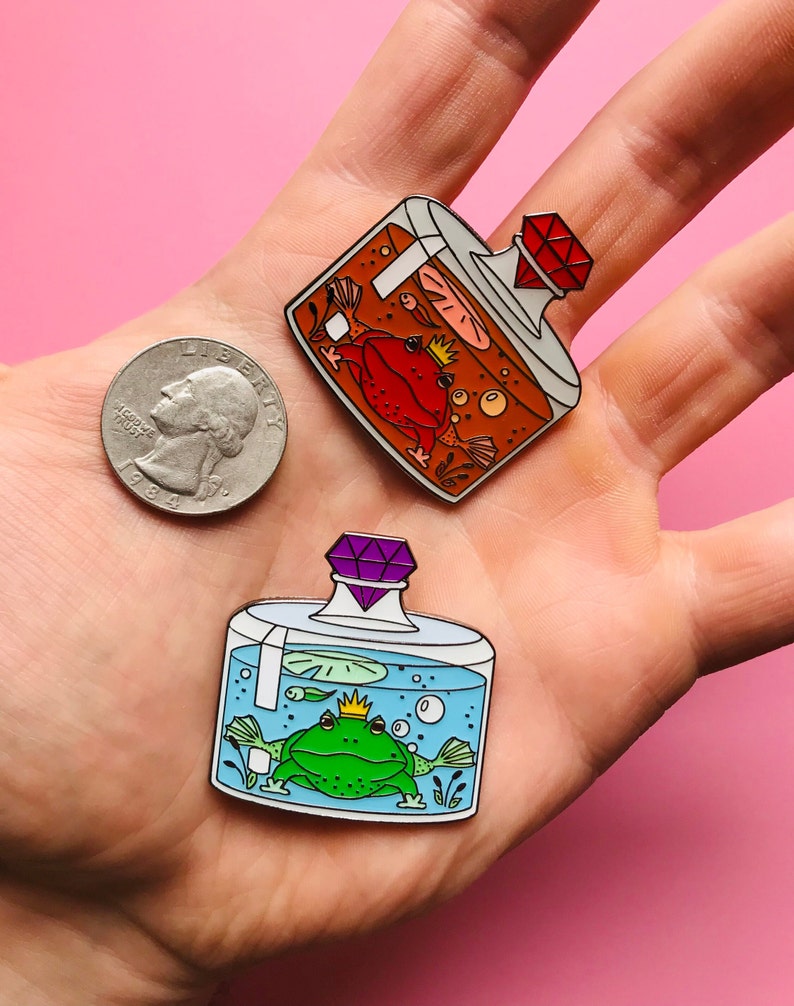 Puede incluir: Dos pines de esmalte que representan ranas en frascos de vidrio con tapas de joyas. El primer pin presenta una rana roja en un frasco naranja con una joya roja. El segundo pin presenta una rana verde en un frasco azul con una joya morada.