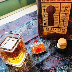 Puede incluir: Un vaso de bourbon whiskey de color &aacute;mbar en un vaso cuadrado con un dise&ntilde;o dorado. Una botella de Kentucky Straight Bourbon Whiskey con la etiqueta "Small Batch 92" est&aacute; en el fondo. Un peque&ntilde;o pin de esmalte de una botella con un cangrejo rojo dentro est&aacute; sobre una tela de estampado paisley azul y blanco.