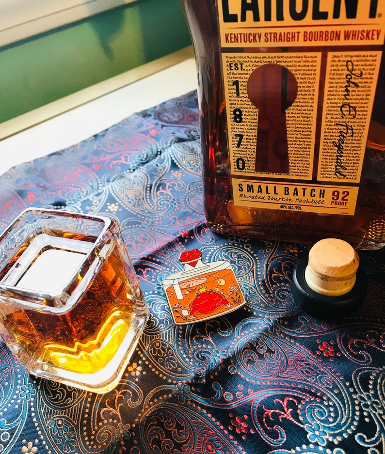 Puede incluir: Un vaso de bourbon whiskey de color &aacute;mbar en un vaso cuadrado con un dise&ntilde;o dorado. Una botella de Kentucky Straight Bourbon Whiskey con la etiqueta "Small Batch 92" est&aacute; en el fondo. Un peque&ntilde;o pin de esmalte de una botella con un cangrejo rojo dentro est&aacute; sobre una tela de estampado paisley azul y blanco.