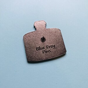 Puede incluir: La parte posterior de un pin de metal plateado con forma de botella con el texto "Blue Frog Pins" en relieve.