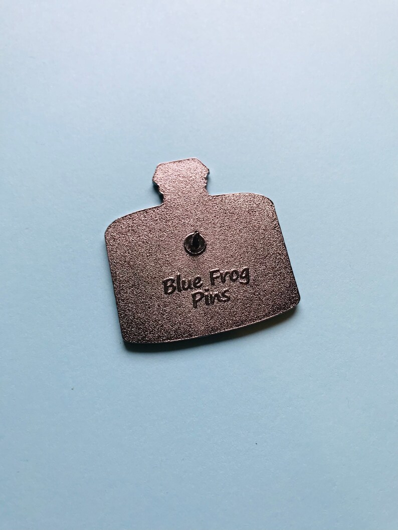 Puede incluir: La parte posterior de un pin de metal plateado con forma de botella con el texto "Blue Frog Pins" en relieve.