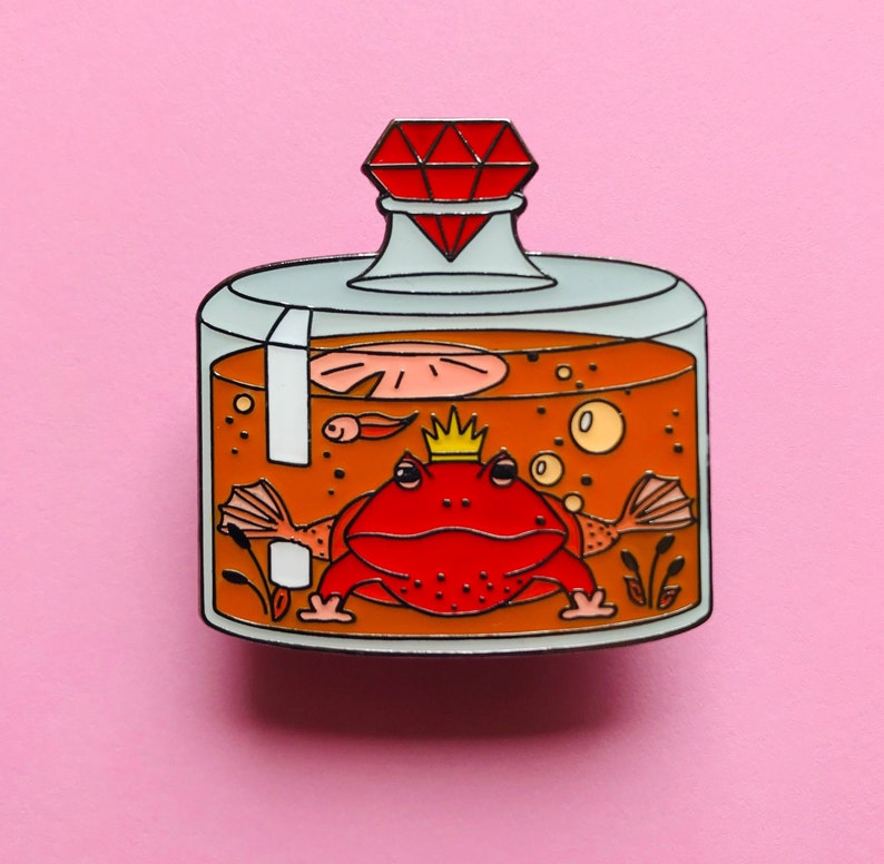 Puede incluir: Una rana roja con una corona dorada se sienta dentro de un frasco de vidrio con un tap&oacute;n de diamante rojo. El frasco est&aacute; lleno de l&iacute;quido naranja y contiene burbujas y un pez peque&ntilde;o.