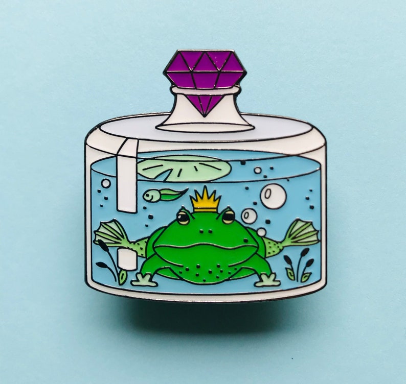 Puede incluir: Un frasco de vidrio con una gema p&uacute;rpura en la parte superior, que contiene una rana verde con una corona dorada. La rana est&aacute; rodeada de agua, burbujas y un nen&uacute;far verde.