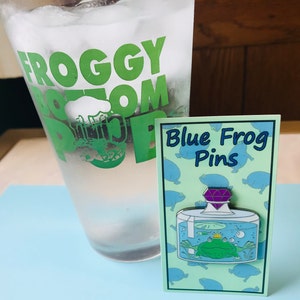 Puede incluir: Un pin de esmalte azul y verde con una rana en un frasco de vidrio con una gema p&uacute;rpura en la parte superior. El pin est&aacute; en una tarjeta con el texto "Blue Frog Pins" y un patr&oacute;n de ranas azules.