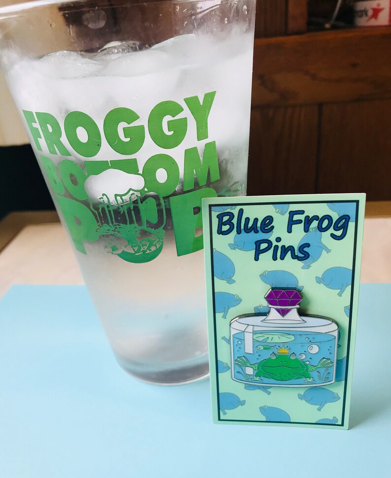 Puede incluir: Un pin de esmalte azul y verde con una rana en un frasco de vidrio con una gema p&uacute;rpura en la parte superior. El pin est&aacute; en una tarjeta con el texto "Blue Frog Pins" y un patr&oacute;n de ranas azules.
