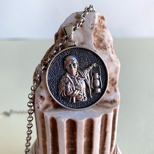K&ouml;nnte beinhalten: Silberfarbene Halskette mit Anh&auml;nger. Der runde Anh&auml;nger zeigt ein detailliertes Relief einer Person, die eine Laterne h&auml;lt. Der Anh&auml;nger h&auml;ngt an einer filigranen Kette. Der Hintergrund ist strukturiert und neutral.
